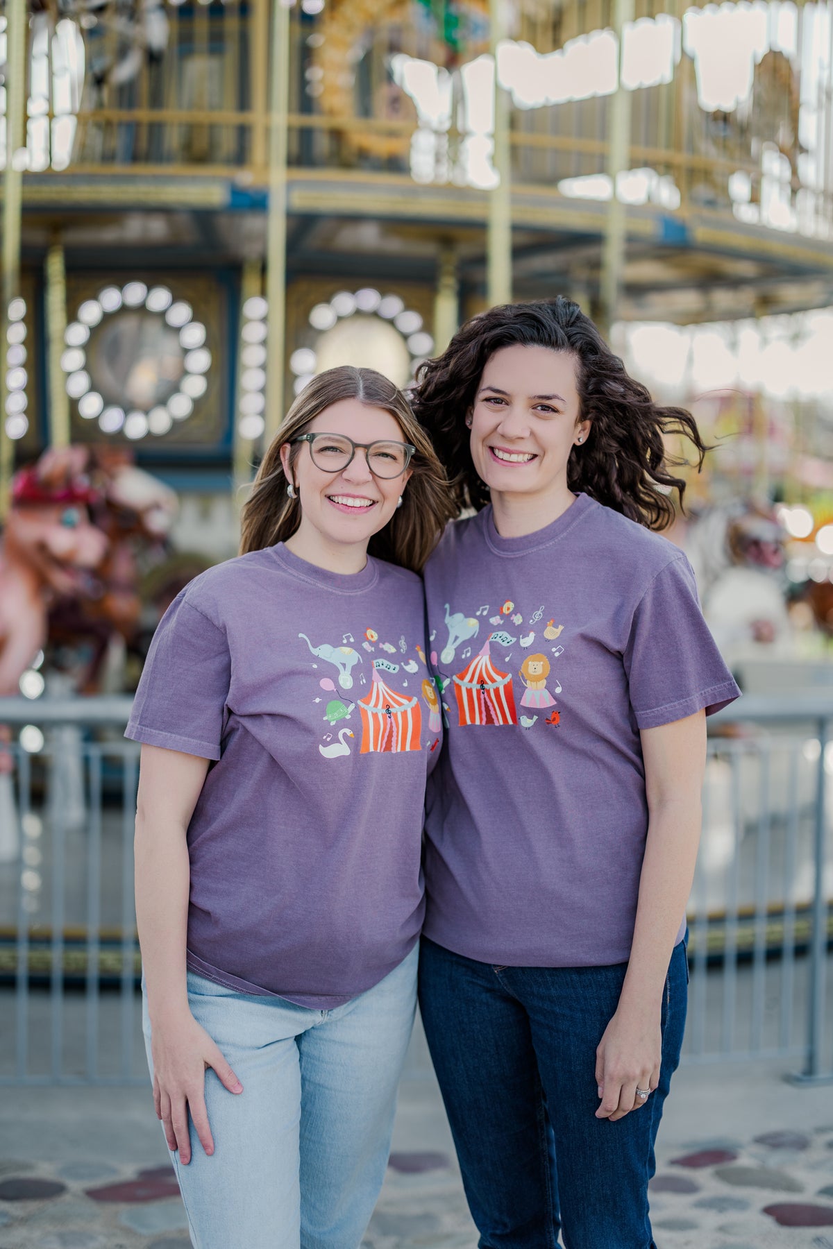 Two people wearing purple t-shirts with a carousel design in front of a merry-go-round.