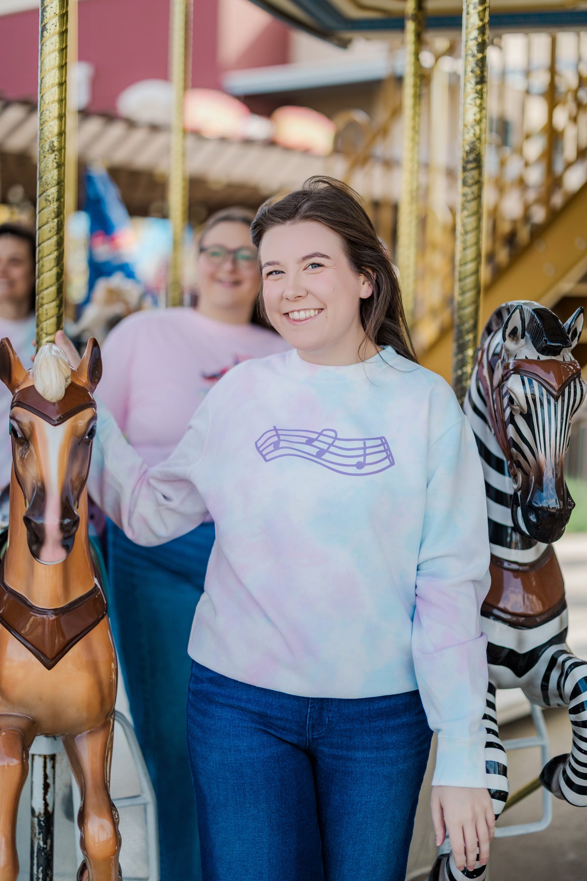 Person wearing a tie-dye sweatshirt with a logo in front of a carousel.