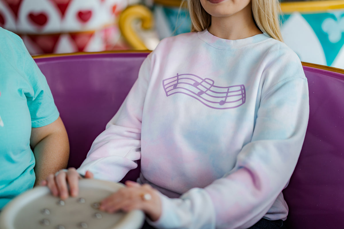 Person wearing a tie-dye sweatshirt with a music note design, sitting on a purple amusement park ride.