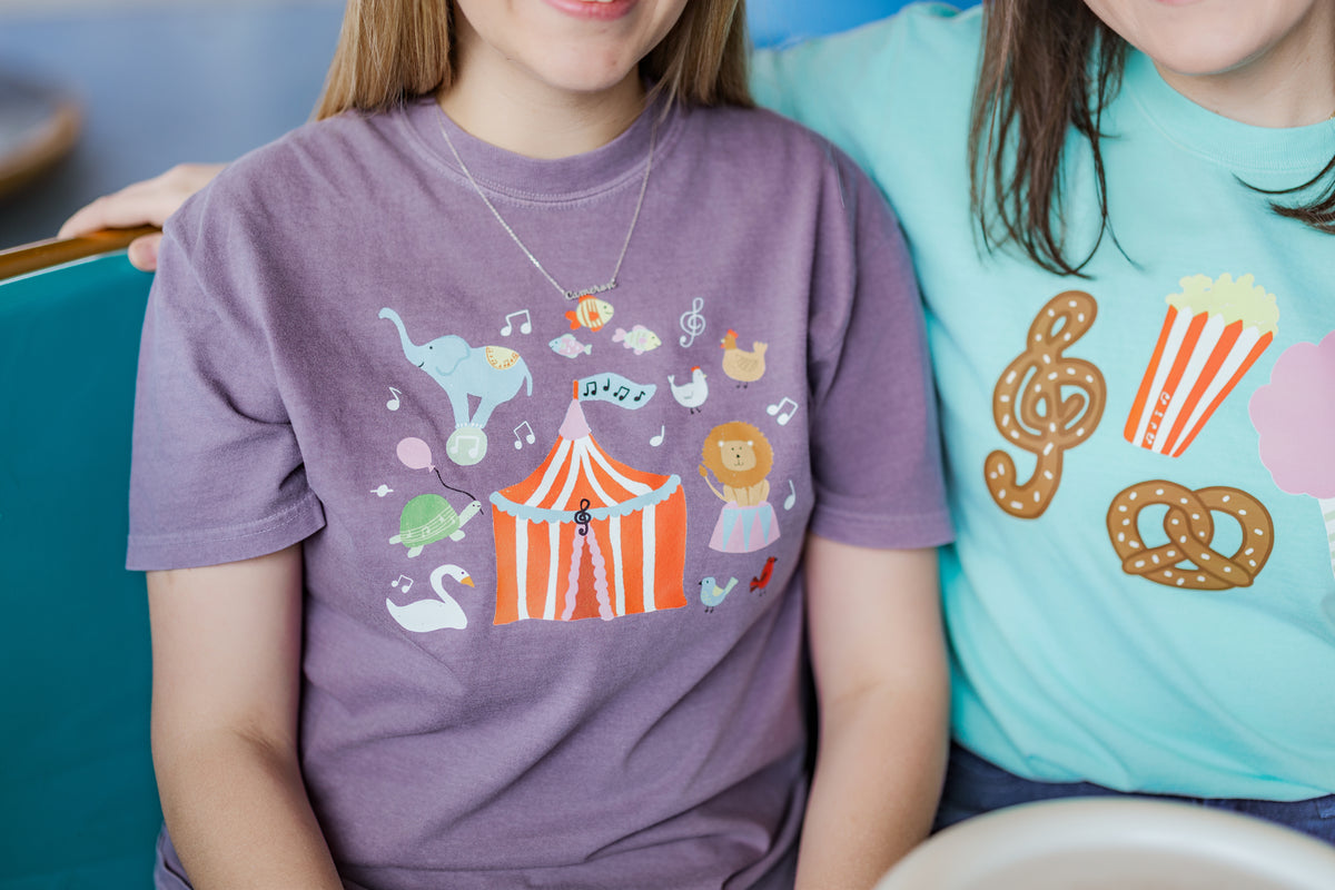 People wearing t-shirts with colorful designs, one purple with a circus-themed design and the other blue with food-themed designs.