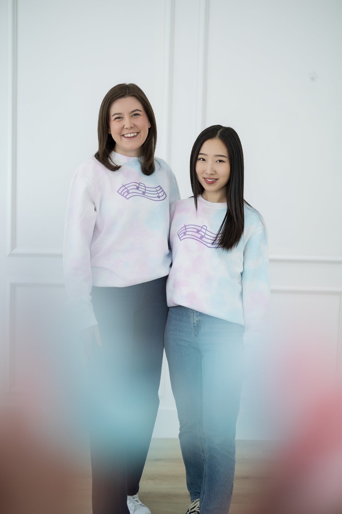 Two women wearing matching light blue and pink sweaters with a music staff, standing against a white wall.