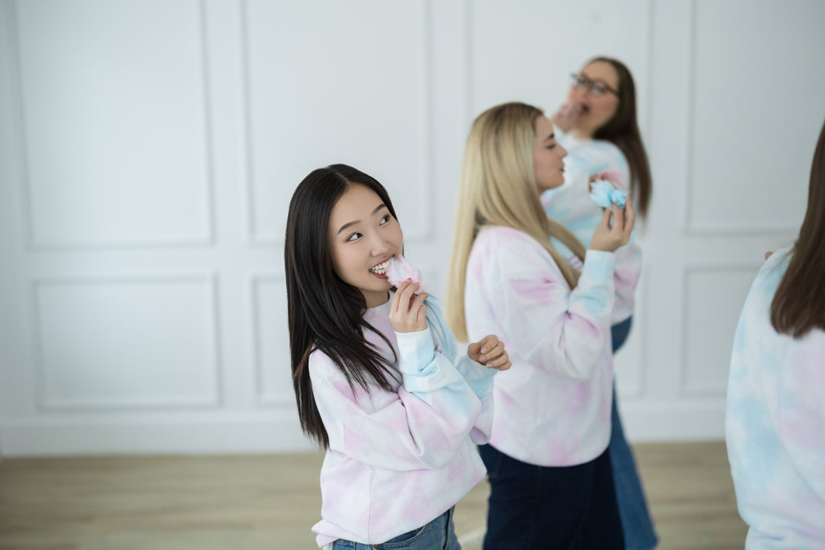 Two women in tie-dye shirts eating cotton candy in a room with white walls.