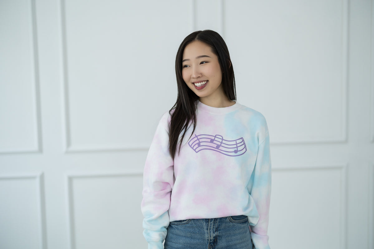 Woman wearing a tie-dye sweatshirt with a graphic design against a white paneled wall.