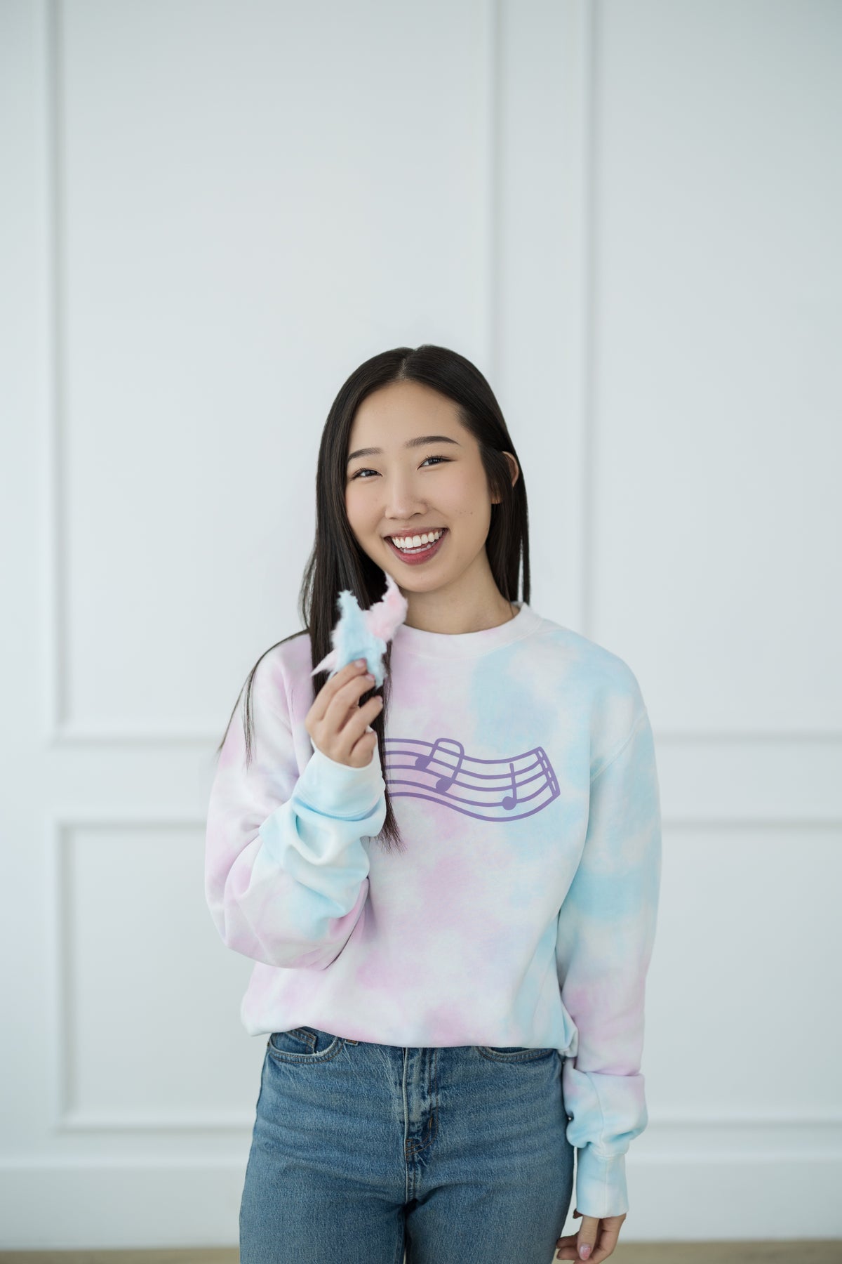 Woman wearing a pastel tie-dye sweater with a graphic design, holding cotton candy against a white paneled wall.