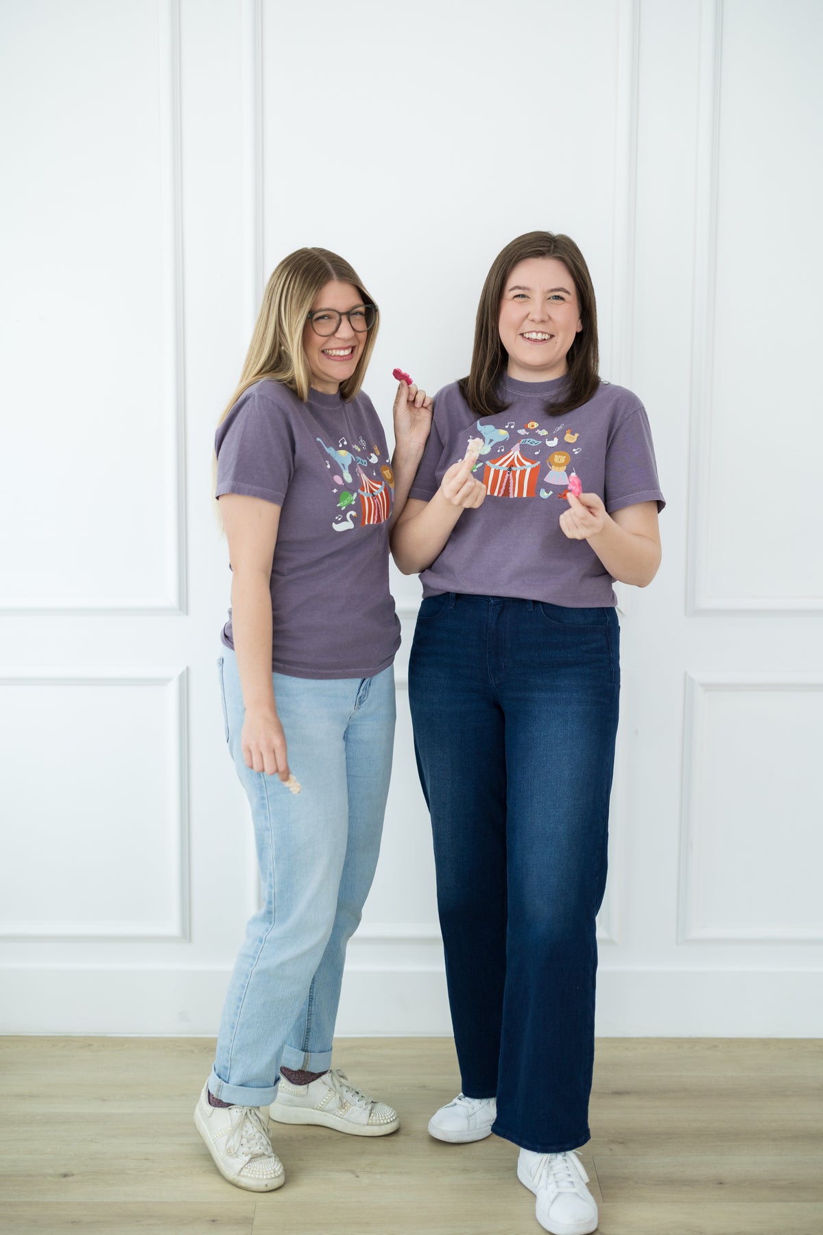 Two women wearing matching purple t-shirts with colorful designs against a white wall.