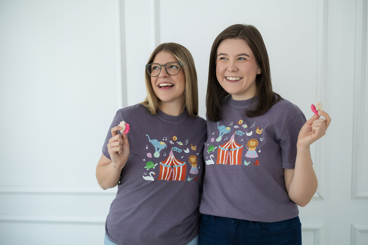 Two women wearing purple t-shirts with colorful designs, holding animal cracker cookies.