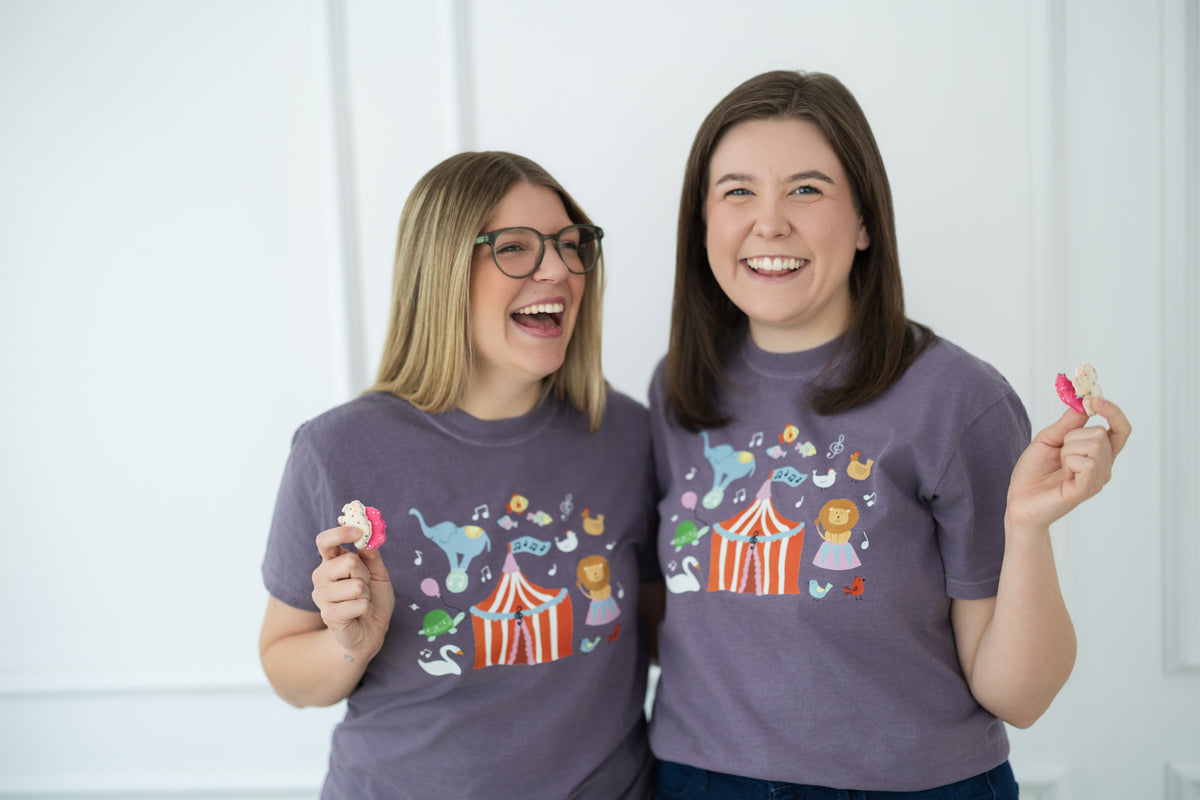 Two women wearing matching purple carnival t-shirts with colorful designs, standing against a white background.