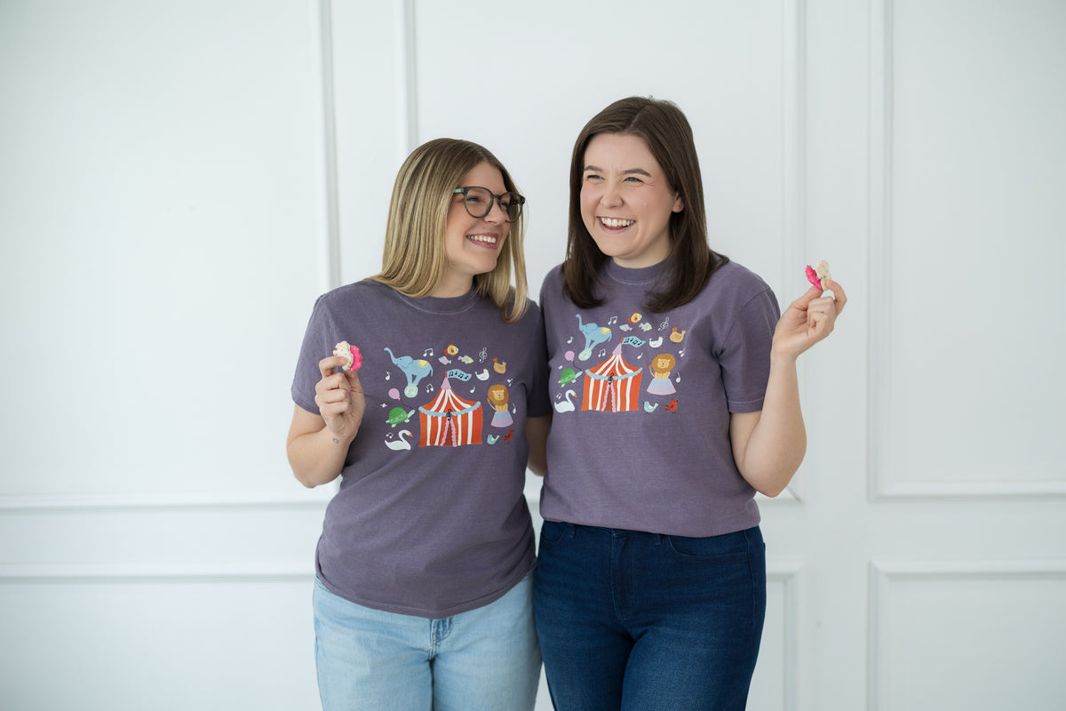 Two women wearing matching purple t-shirts with a colorful design, standing against a white wall.
