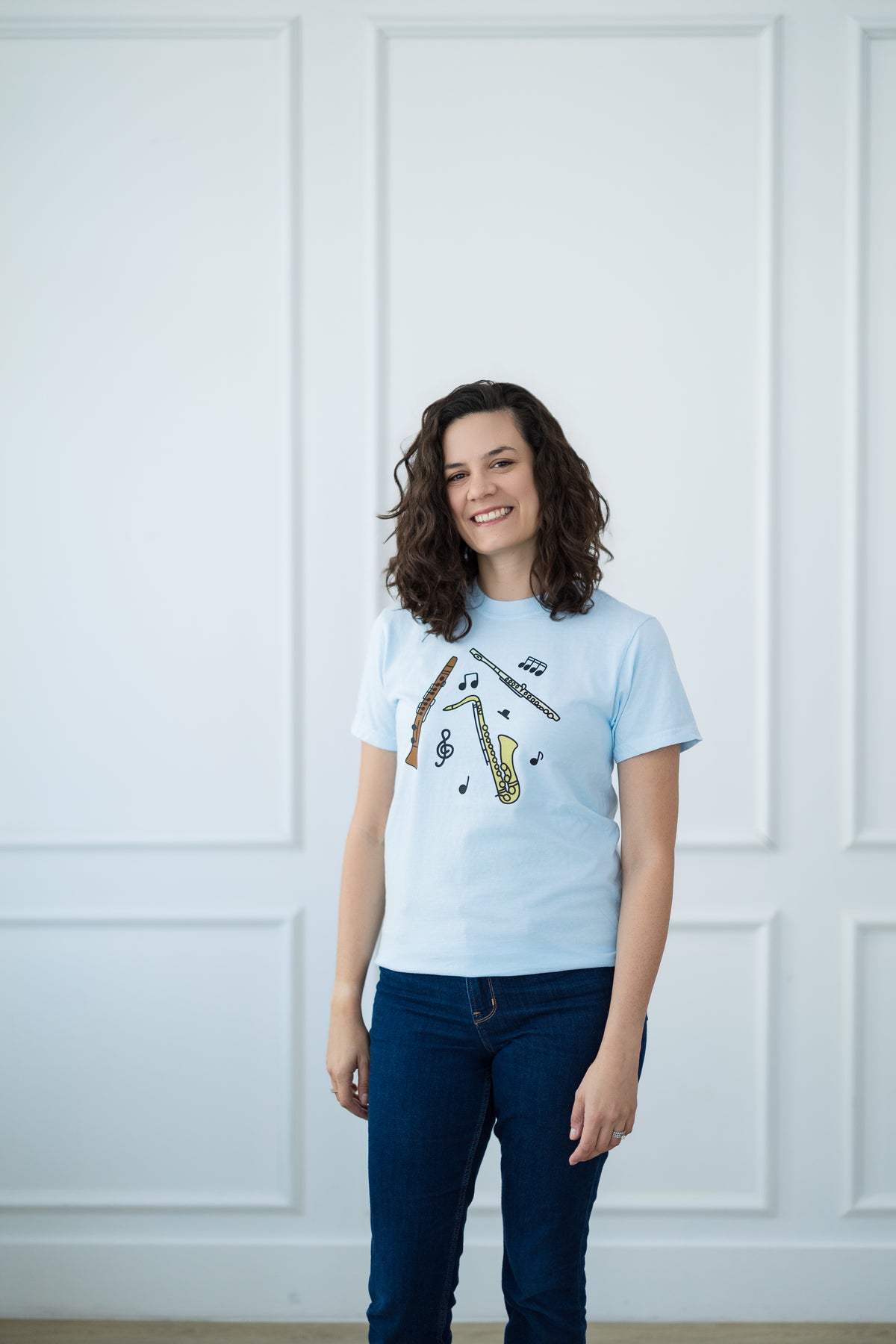Woman wearing a light blue t-shirt with a graphic design against a white paneled wall.