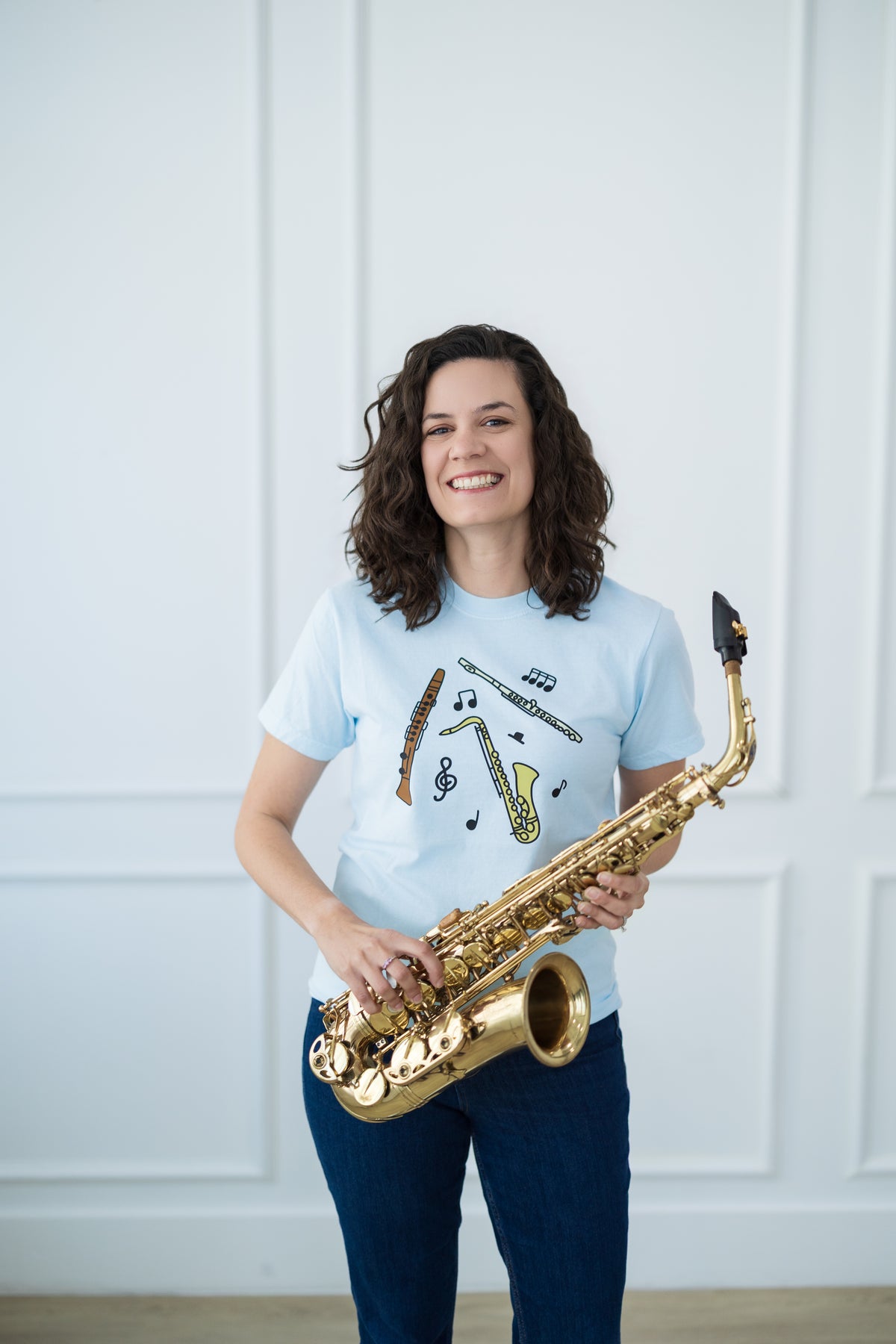 Person holding a saxophone wearing a light blue t-shirt with musical designs against a white paneled wall.