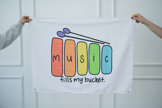 White tapestry with colorful 'Music fills my bucket' design held by two people against a white background