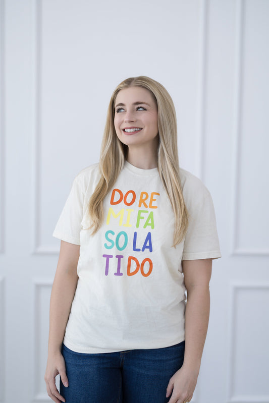 Woman wearing a white t-shirt with colorful text against a white paneled wall.