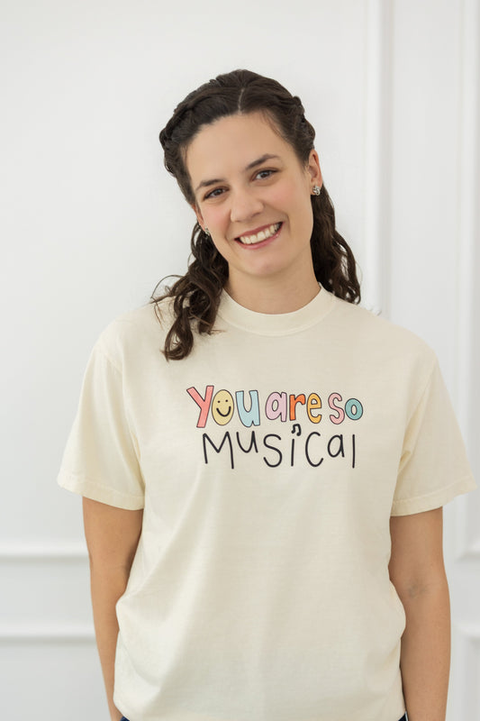 Person wearing a cream t-shirt with 'You are so musical' text, standing against a white paneled wall.