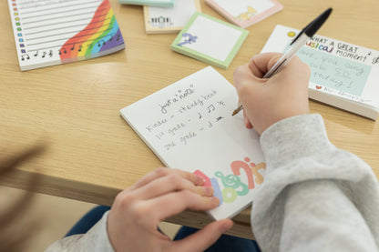 Person writing in a small notebook with colorful stationery on a wooden table.
