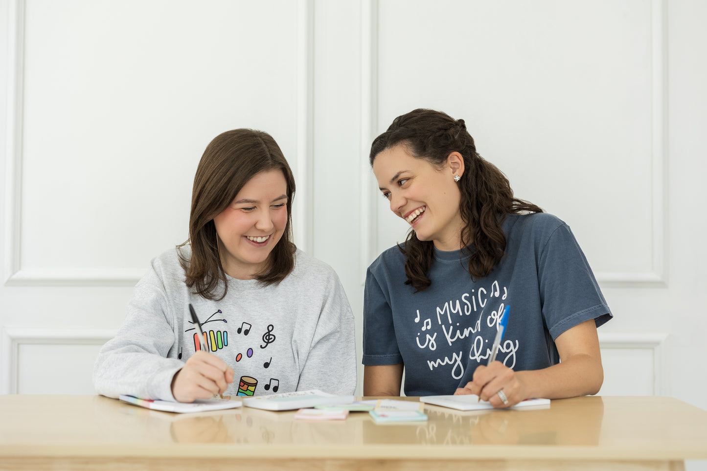 Two women sitting at a table with notebooks, smiling and engaged in a conversation.