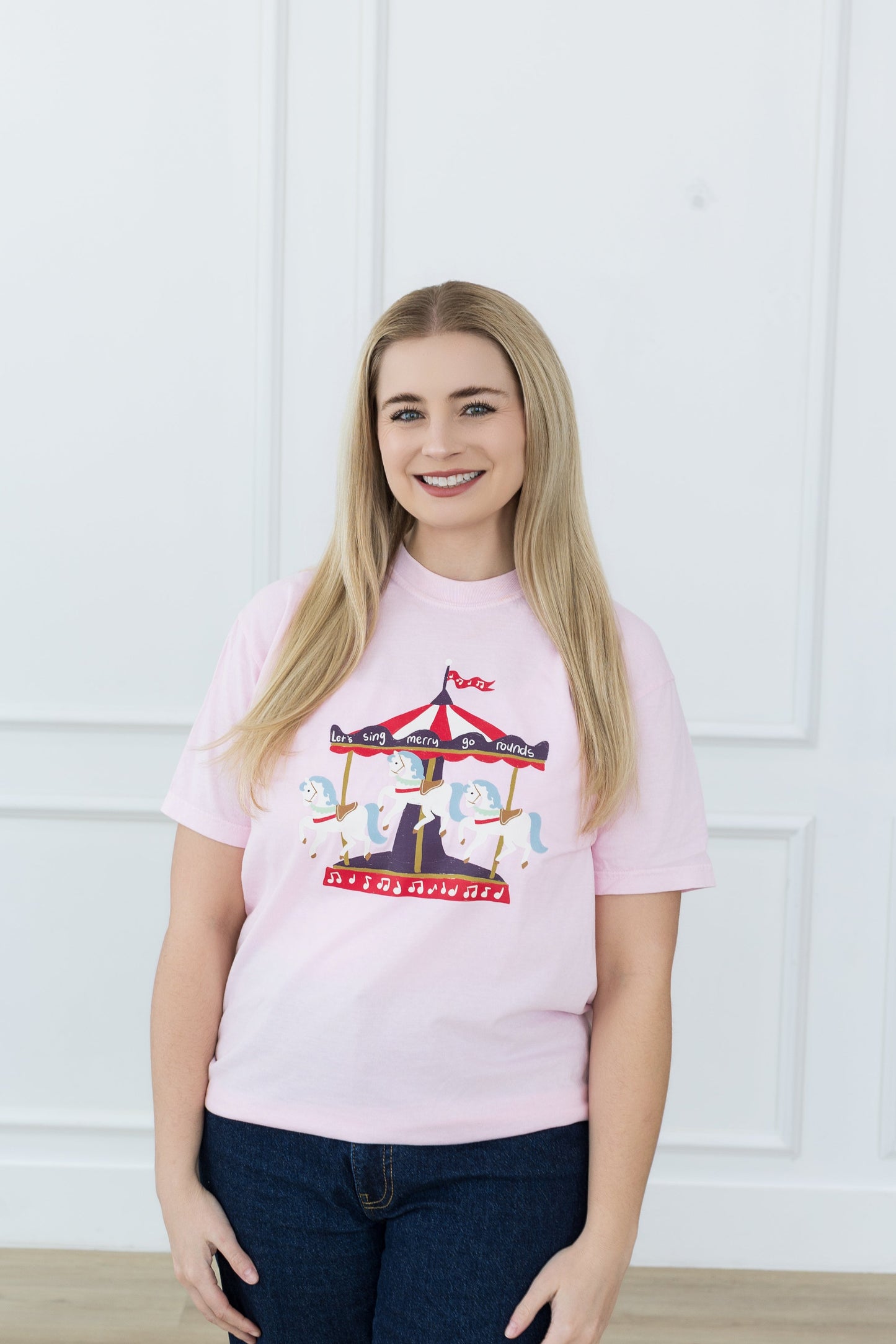 Woman wearing a pink t-shirt with a graphic design against a white paneled wall.