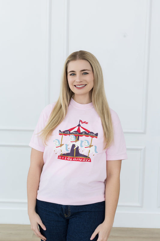 Woman wearing a pink t-shirt with a graphic design against a white paneled wall.