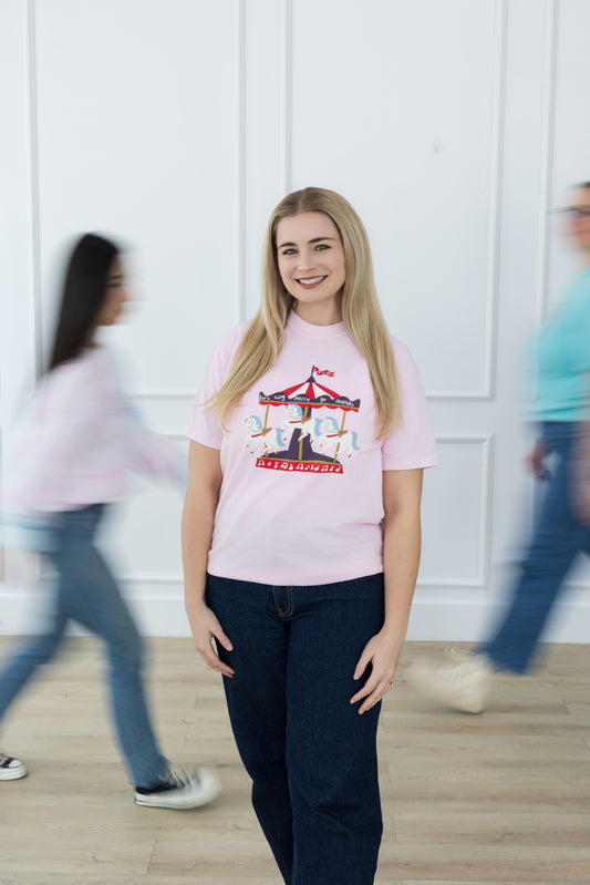 Woman wearing a pink t-shirt with a graphic design, standing in a room with blurred people in the background.
