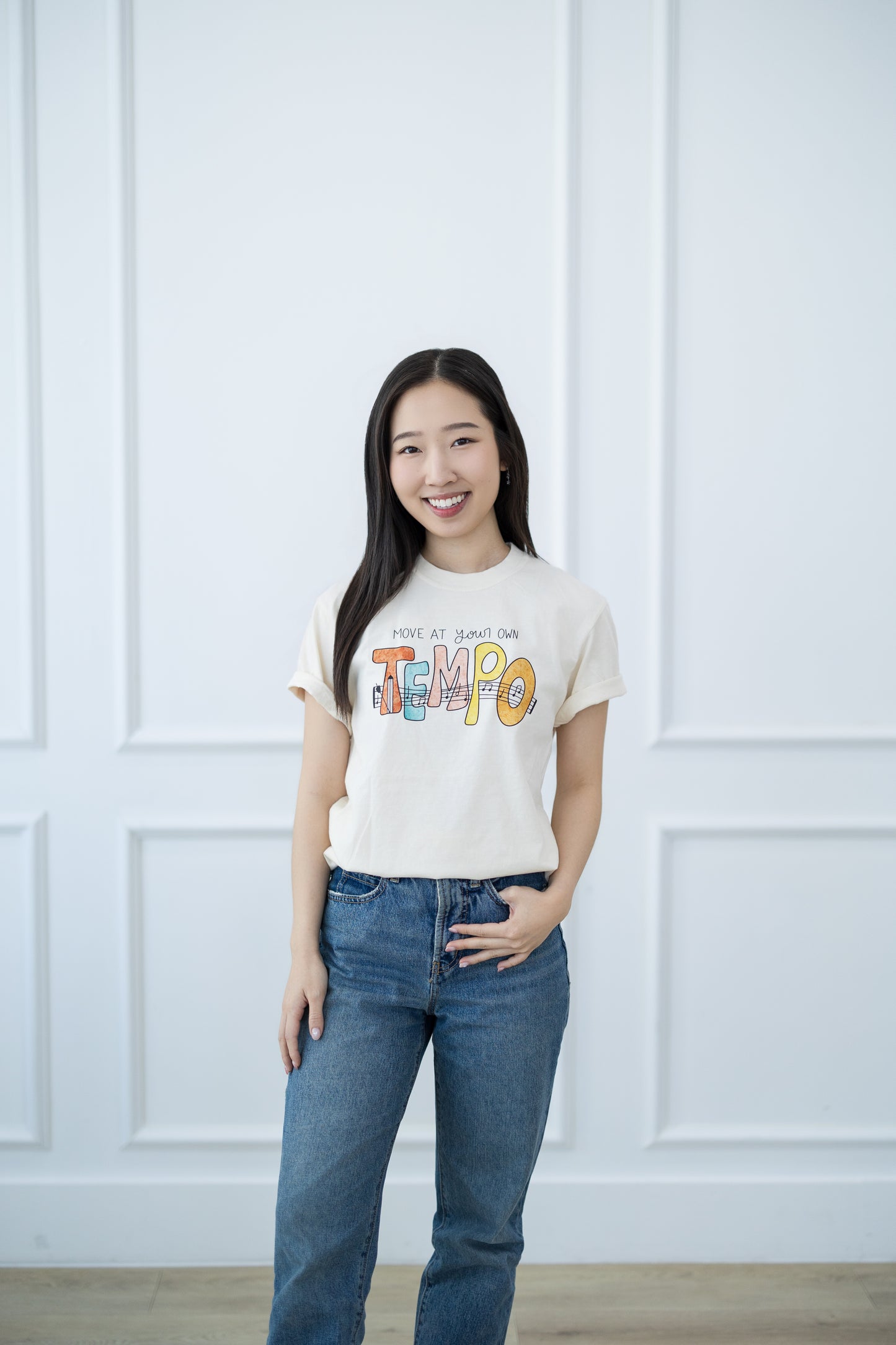 Woman wearing a white t-shirt with colorful text and blue jeans against a white paneled wall.