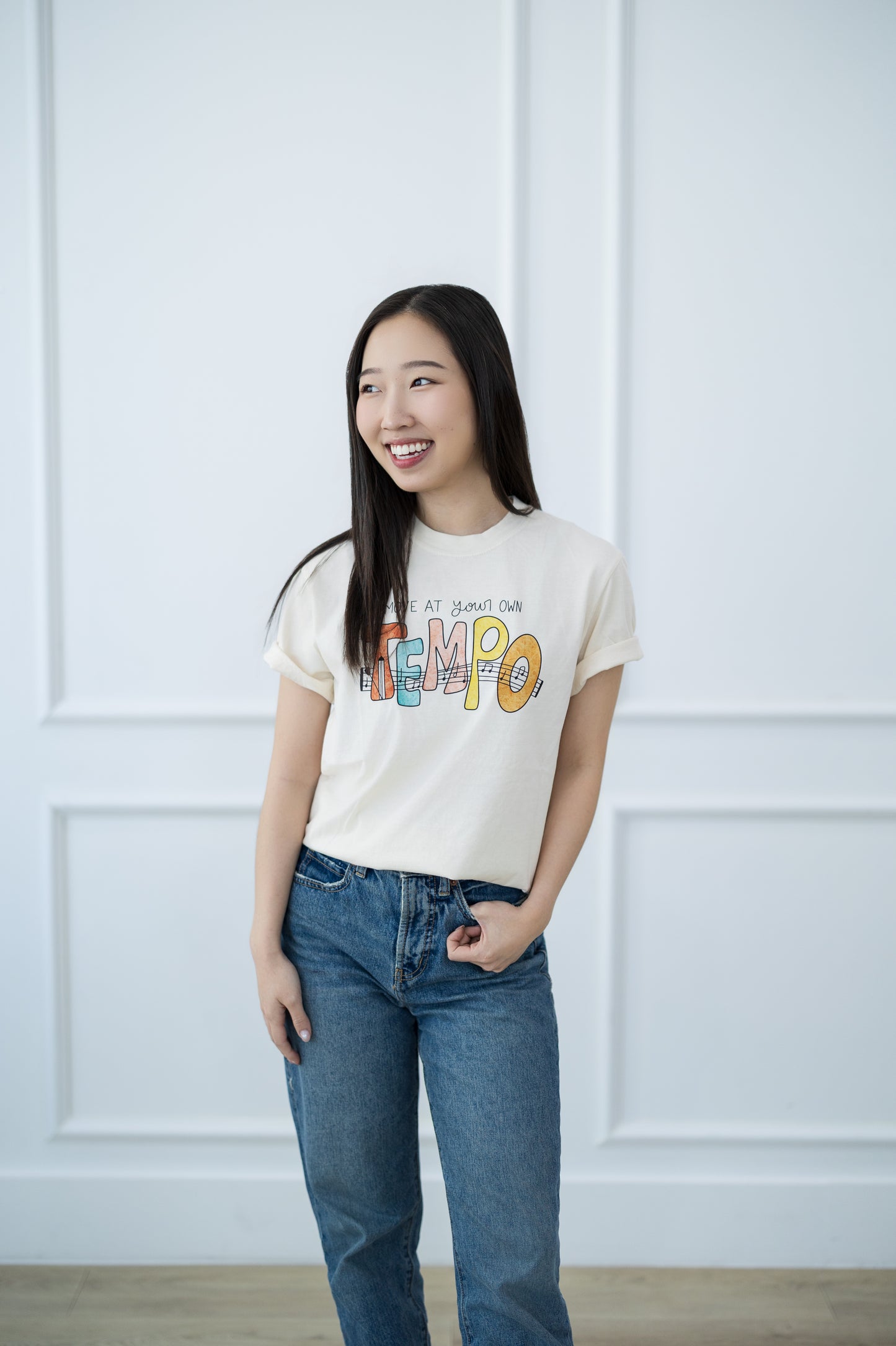 Woman wearing a white t-shirt with colorful text and blue jeans against a white paneled wall.