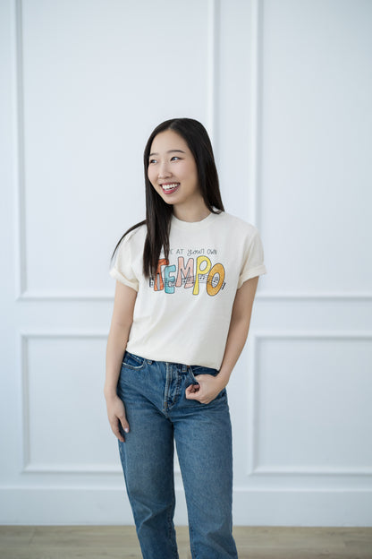 Woman wearing a white t-shirt with colorful text and blue jeans against a white paneled wall.