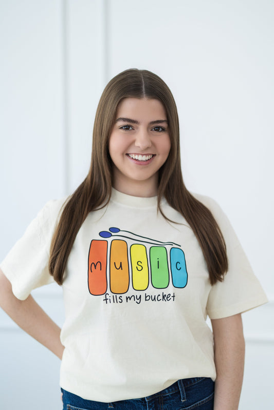 Woman wearing a white t-shirt with colorful 'Music fills my bucket!' design against a light background