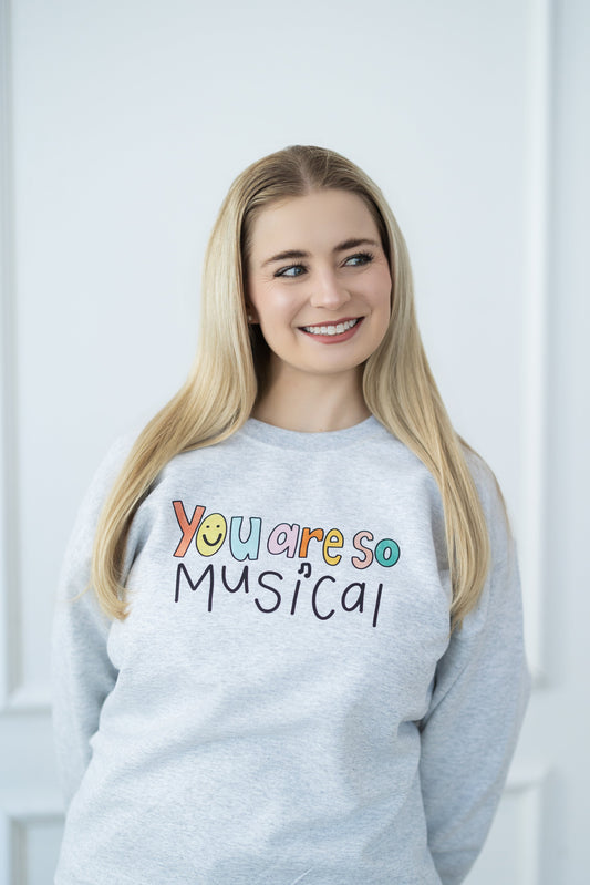 Woman wearing a gray sweatshirt with 'You are so musical!' text, standing against a white wall.