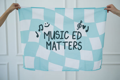 Checkered tapestry with 'Music Ed Matters' text held by two hands against a white background