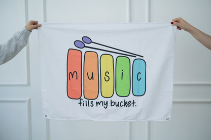 White tapestry with colorful 'Music fills my bucket' design held by two people against a white background