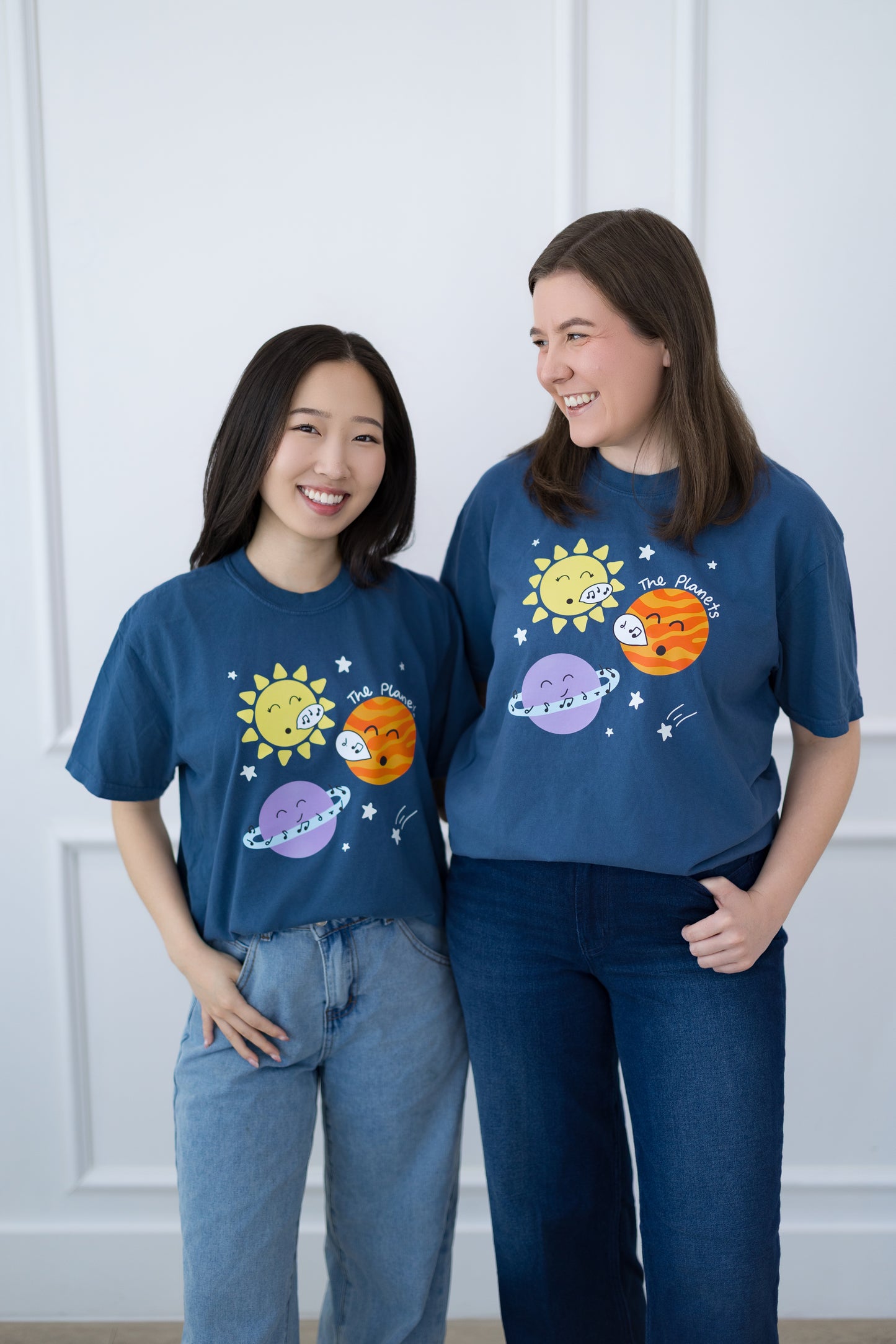Two people wearing matching blue t-shirts with solar system designs against a white paneled wall.