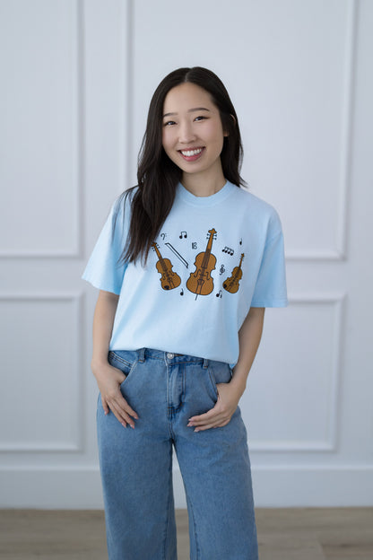 Woman wearing a light blue t-shirt with musical instrument design against a white paneled wall.