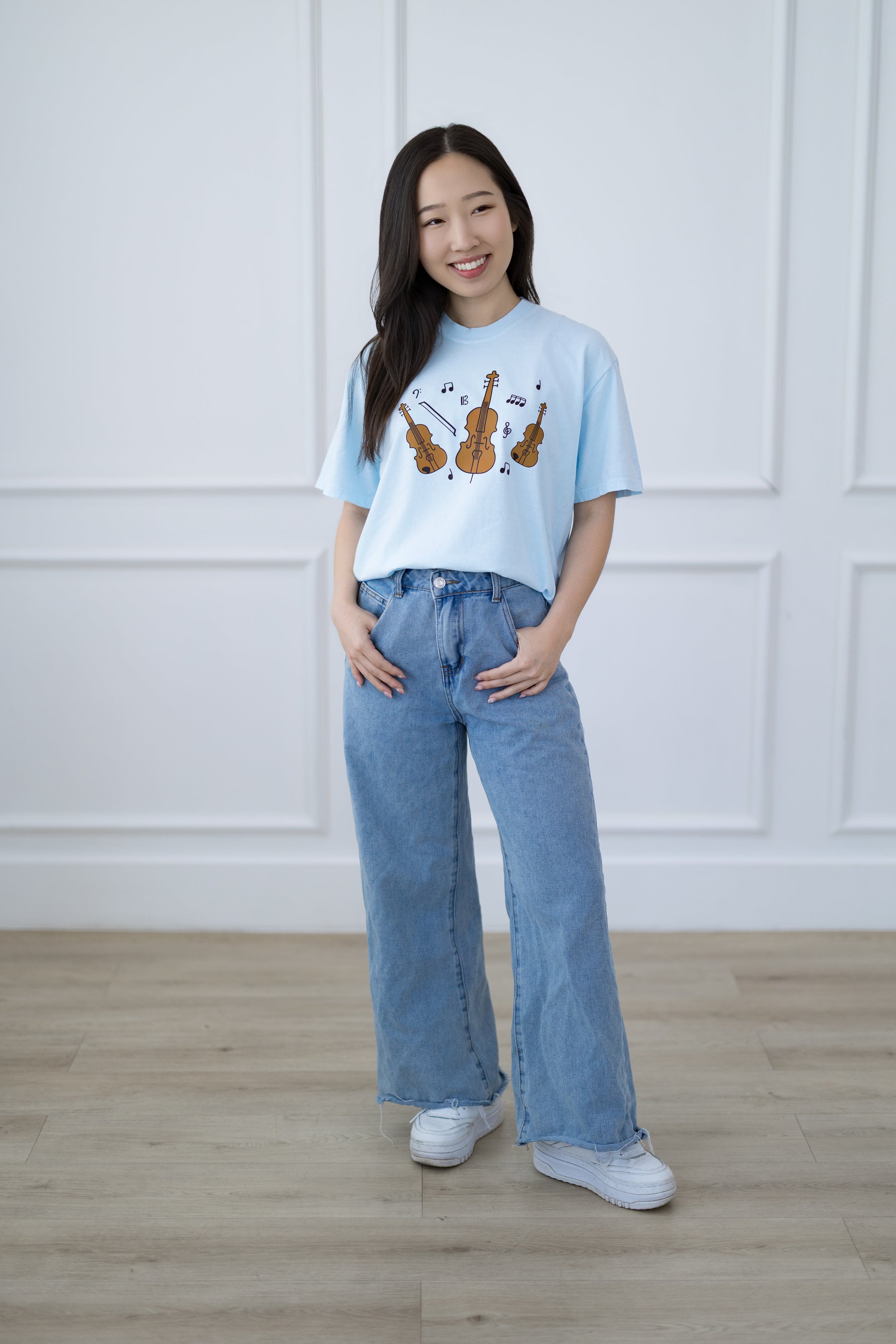 Woman wearing a light blue t-shirt with string instrument designs and blue jeans against a white paneled wall.