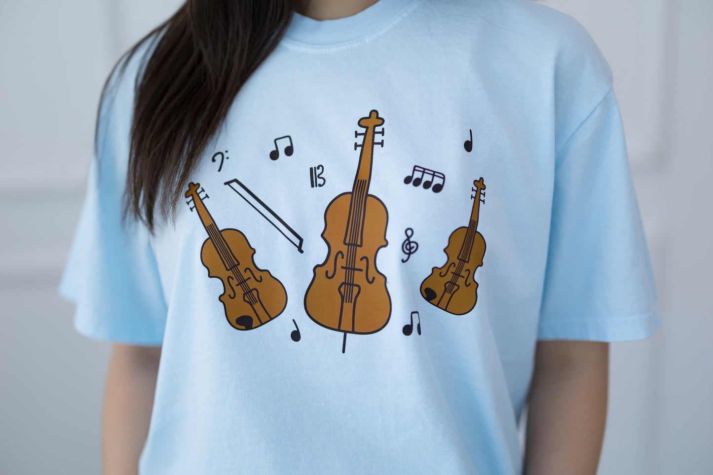 Light blue t-shirt with brown violin and cello illustrations and music notes on a plain background
