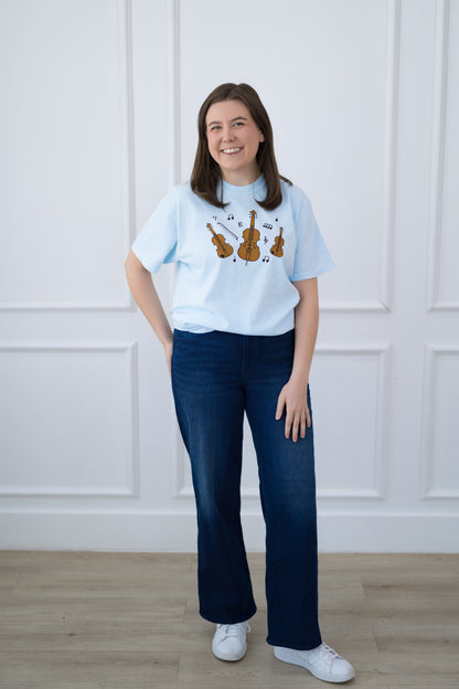 Person wearing a light blue t-shirt with graphic design and dark blue jeans against a white paneled wall.