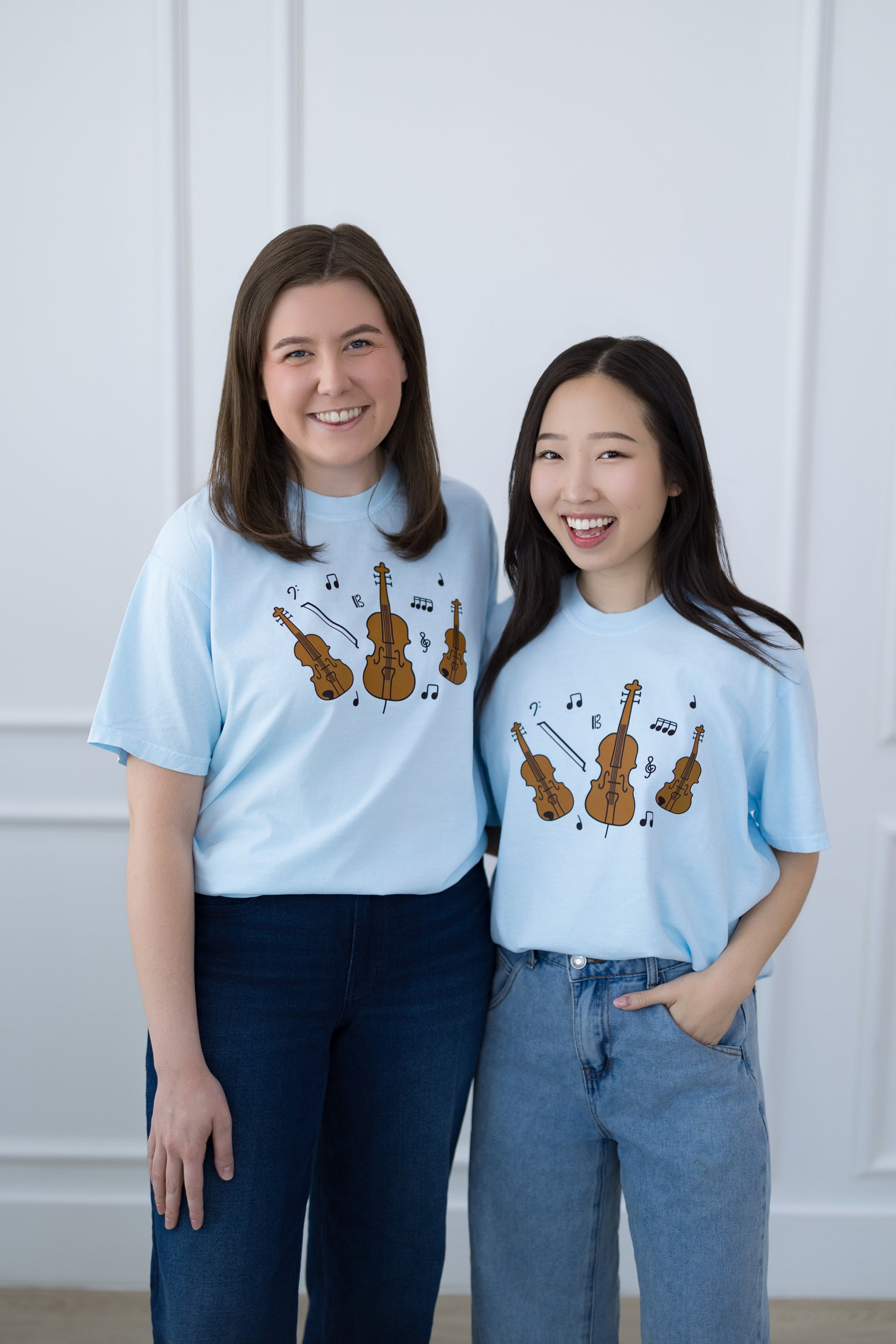 Two women wearing light blue t-shirts with musical instrument designs against a white wall.
