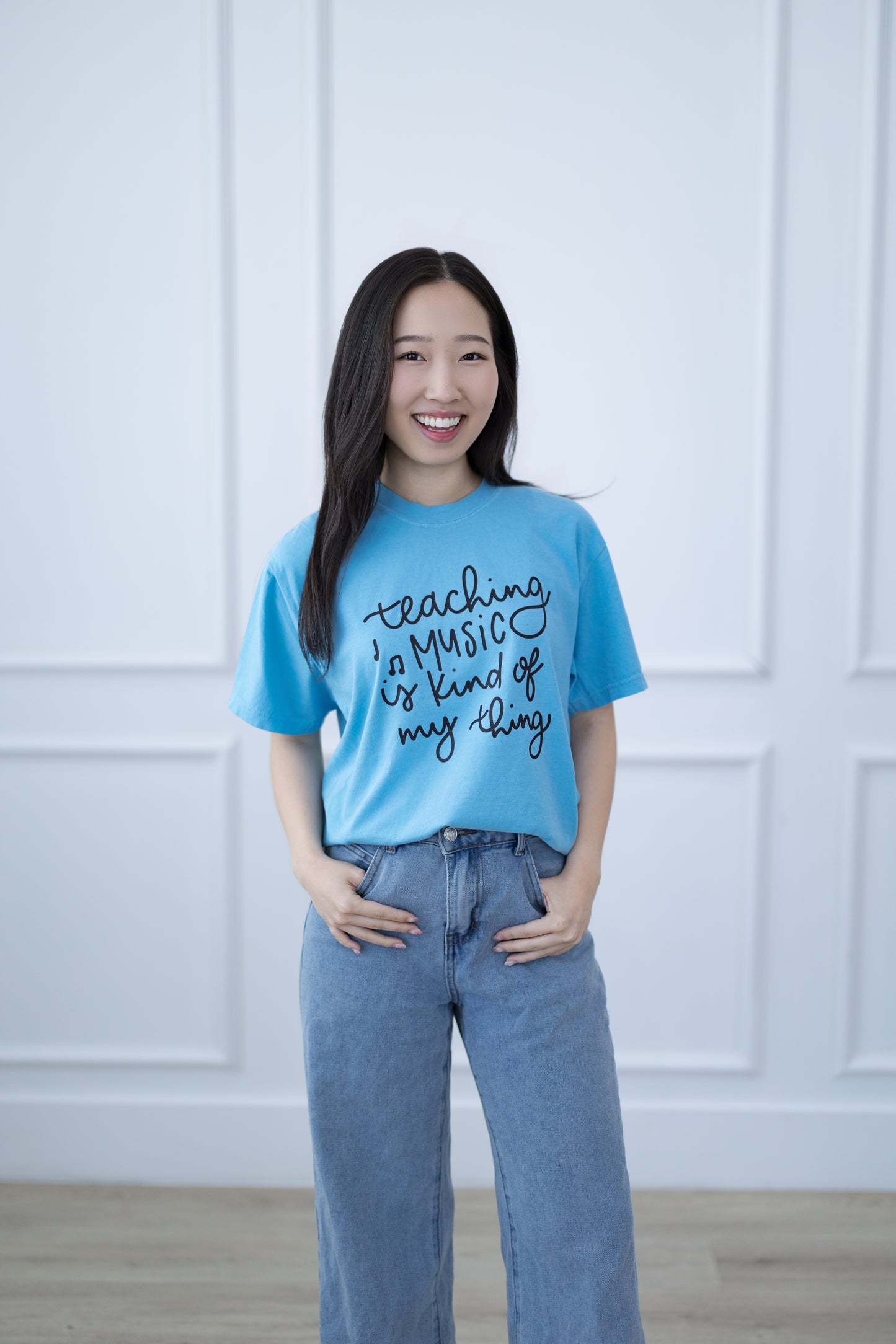 Woman wearing a blue t-shirt with text, standing against a white paneled wall.