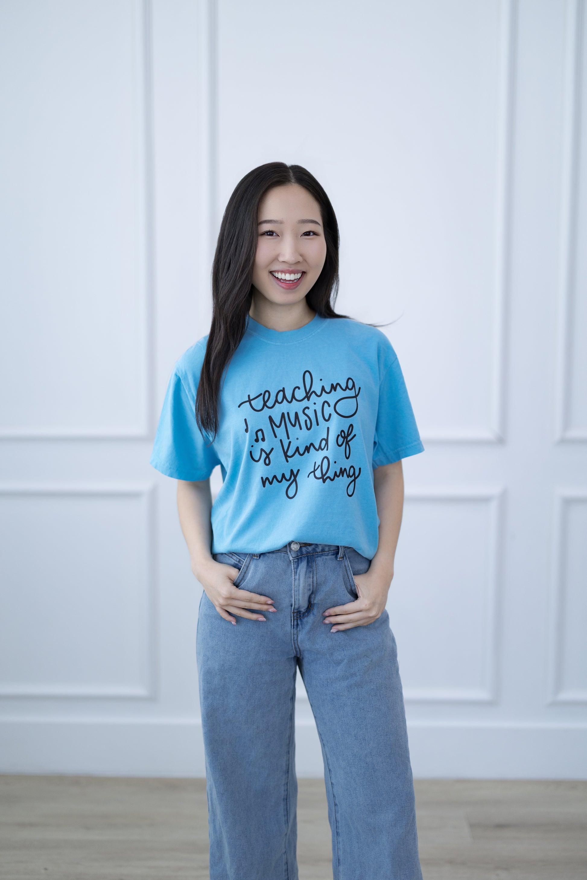 Woman wearing a blue t-shirt with text, standing against a white paneled wall.