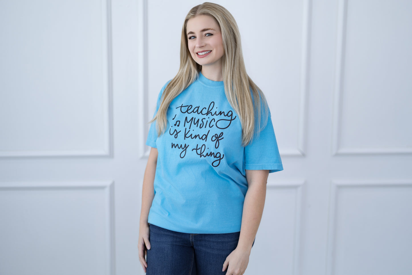 Woman wearing a blue t-shirt with text, standing against a white paneled wall.