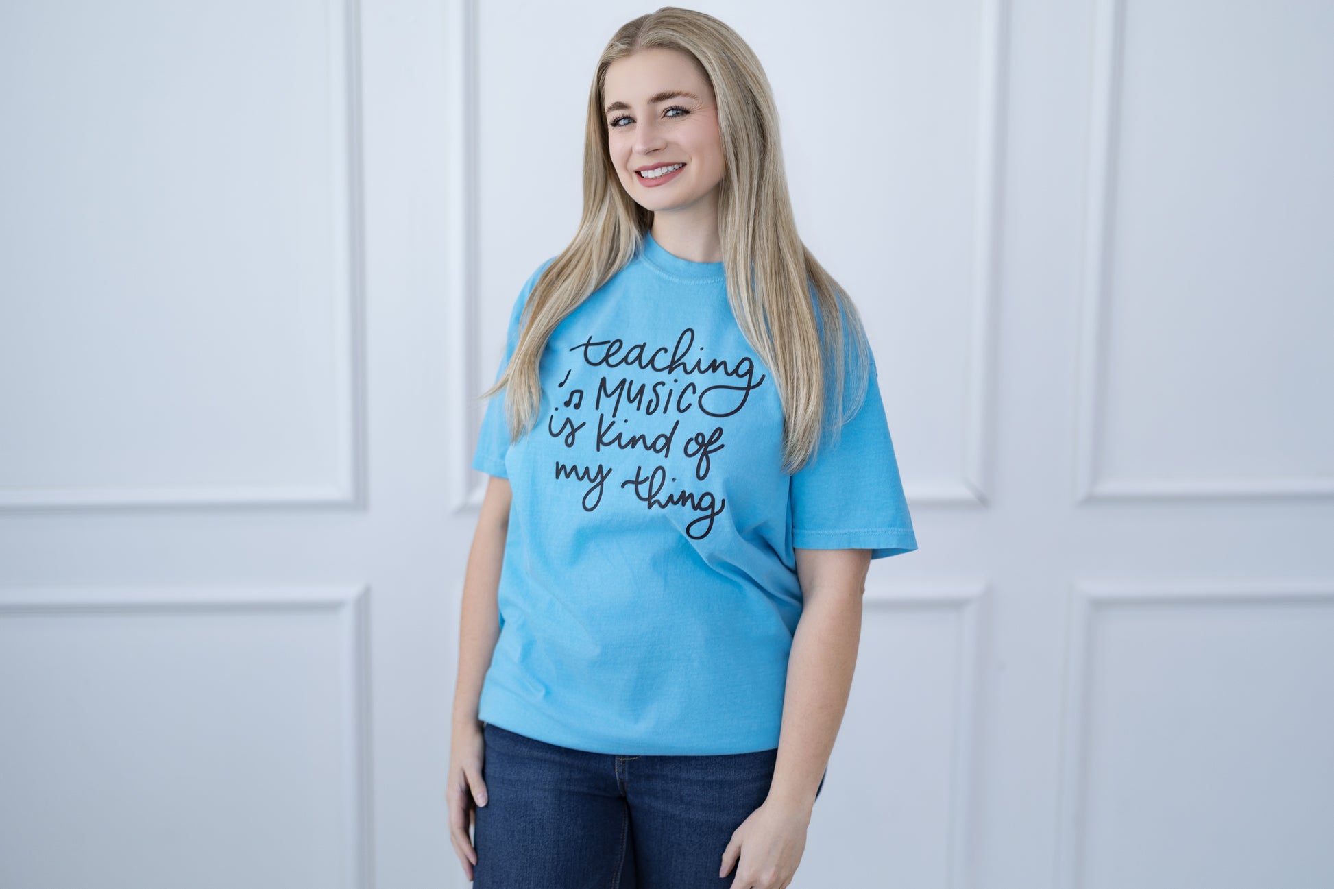 Woman wearing a blue t-shirt with text, standing against a white paneled wall.