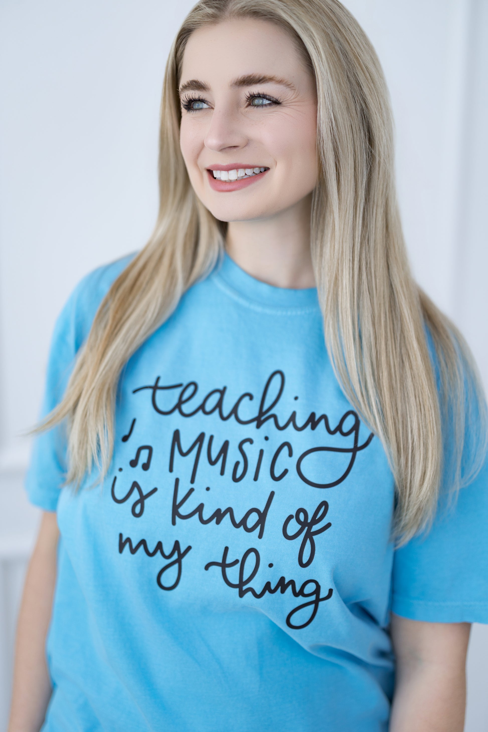 Person wearing a blue t-shirt with text 'teaching music is kind of my thing' on a plain background