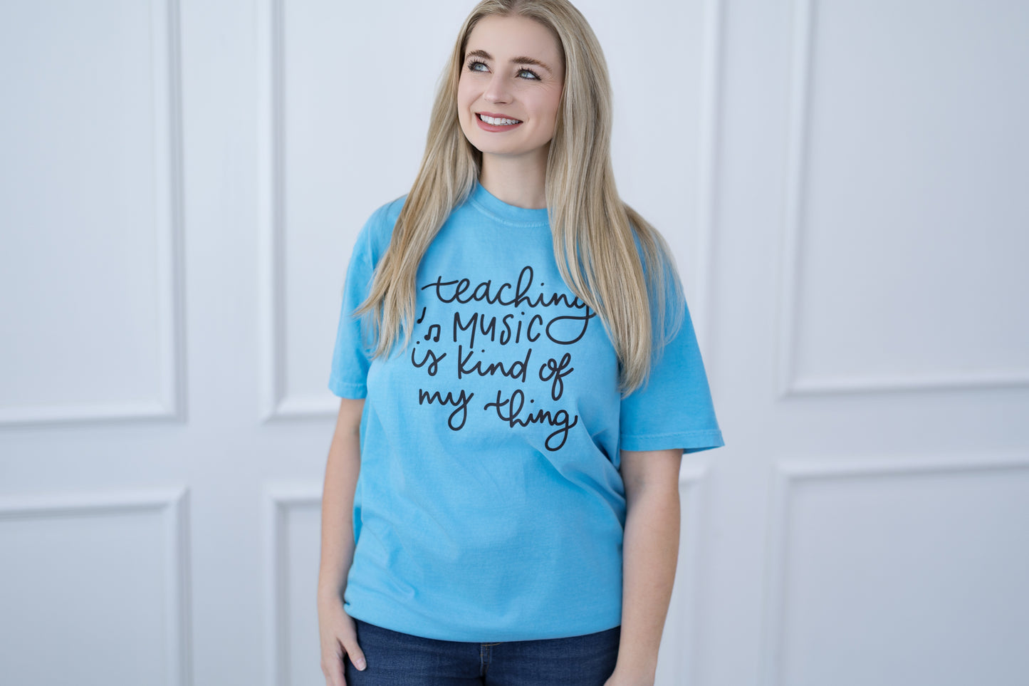 Person wearing a blue t-shirt with 'teaching music is kind of my thing' text, standing against a white paneled wall.