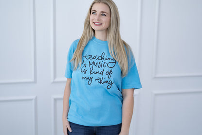 Person wearing a blue t-shirt with 'teaching music is kind of my thing' text, standing against a white paneled wall.