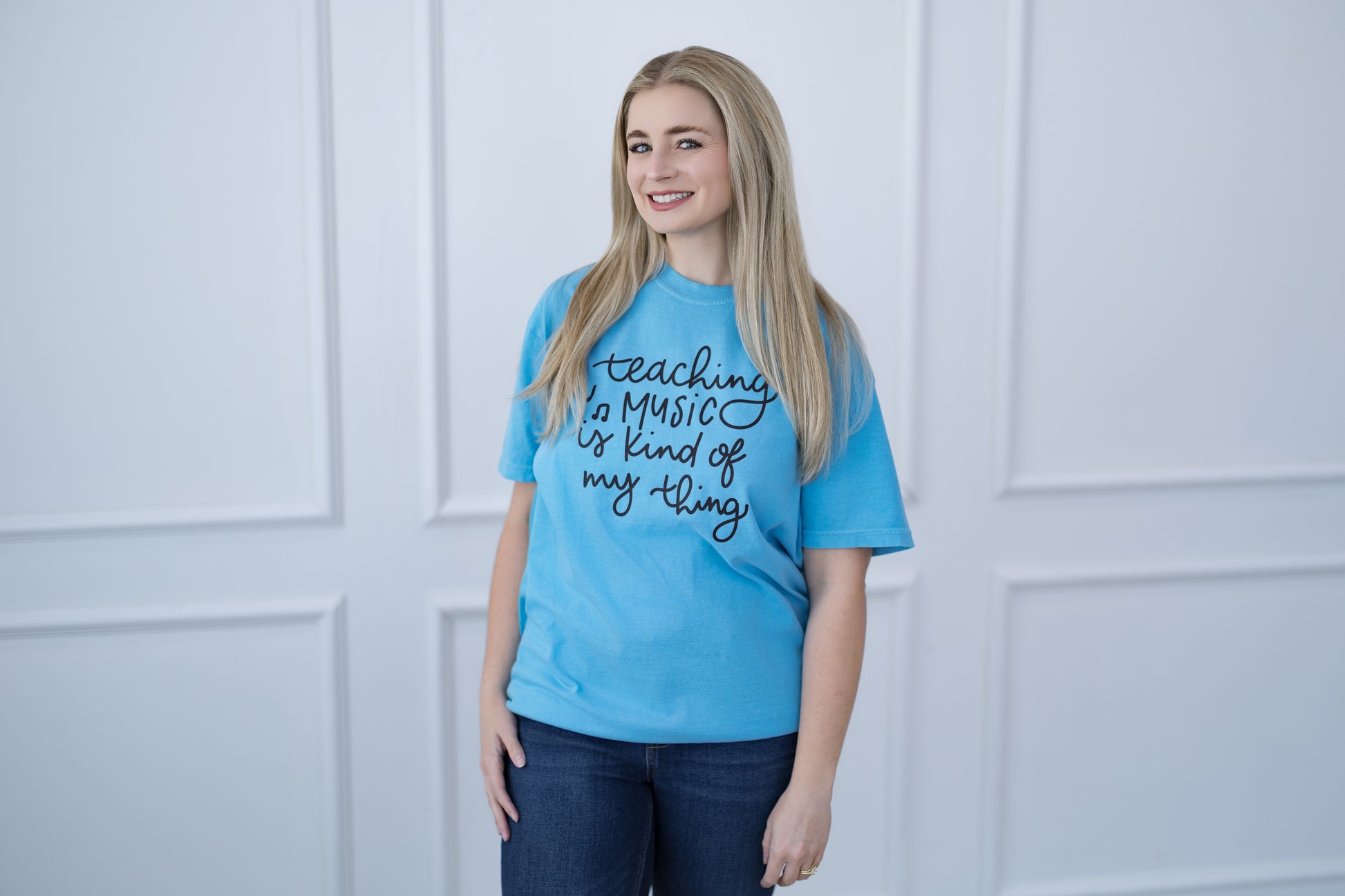 Woman wearing a blue t-shirt with text, standing against a white paneled wall.