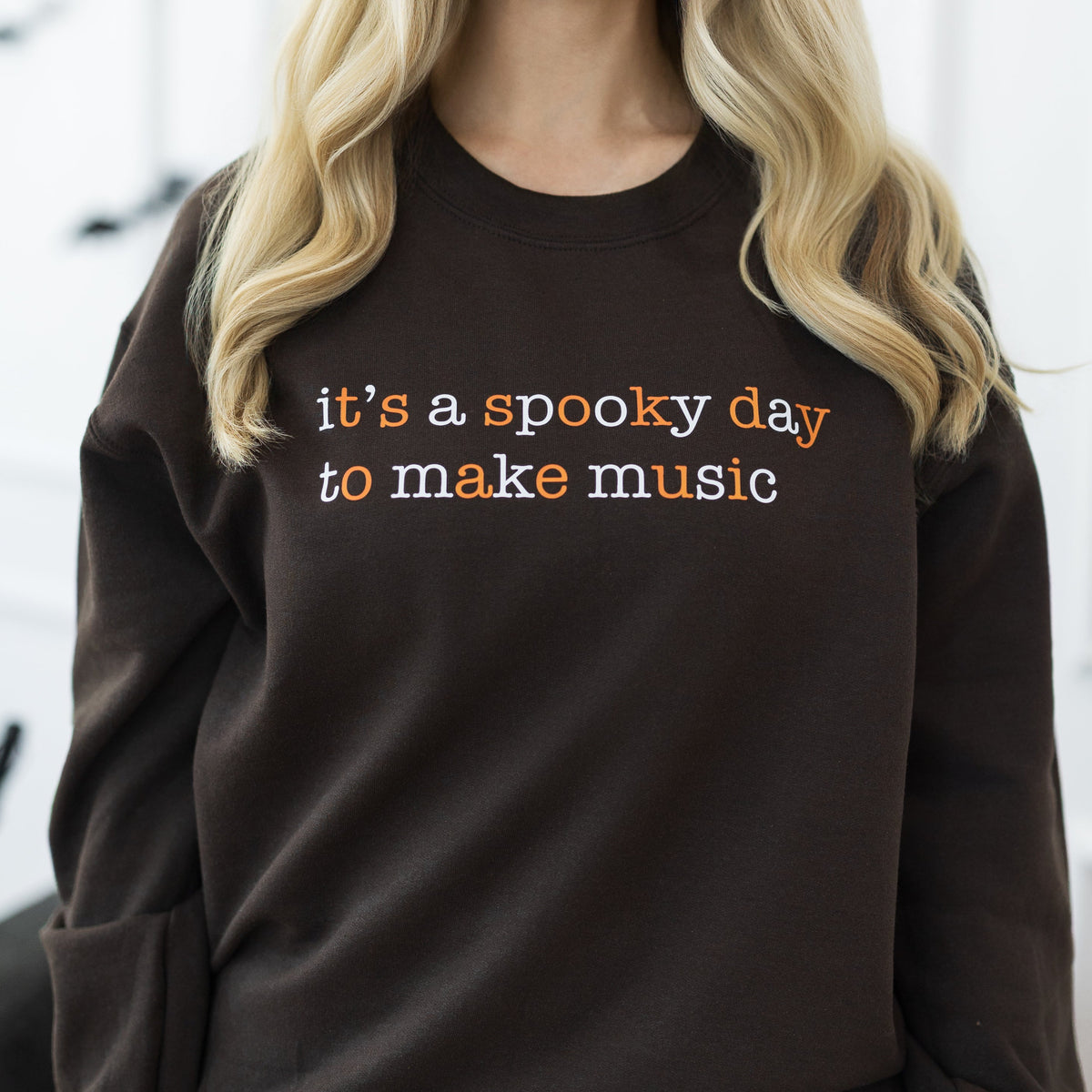 A woman with long blonde hair wearing a dark brown crewneck sweatshirt with the text 'it's a spooky day to make music' in orange and white, paired with dark blue jeans. She is standing in a decorated indoor setting with Halloween elements like bats on the wall and a pumpkin on the floor.