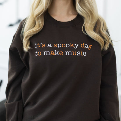 A woman with long blonde hair wearing a dark brown crewneck sweatshirt with the text 'it's a spooky day to make music' in orange and white, paired with dark blue jeans. She is standing in a decorated indoor setting with Halloween elements like bats on the wall and a pumpkin on the floor.