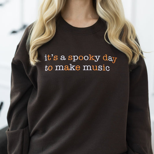 A woman with long blonde hair wearing a dark brown crewneck sweatshirt with the text 'it's a spooky day to make music' in orange and white, paired with dark blue jeans. She is standing in a decorated indoor setting with Halloween elements like bats on the wall and a pumpkin on the floor.