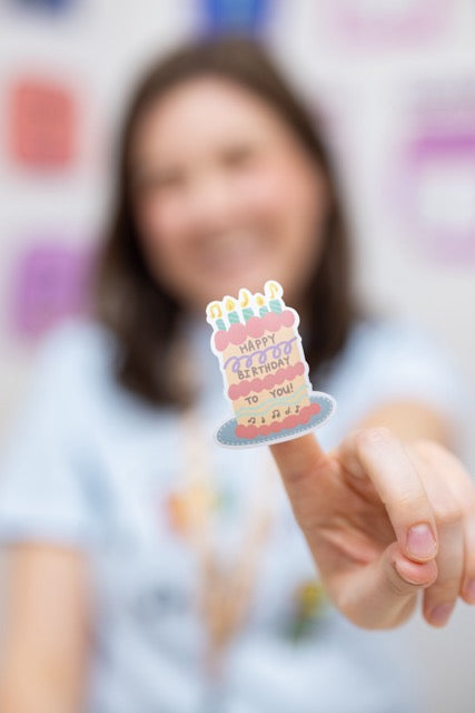 Person holding a small birthday cake sticker with colorful candles against a blurred background