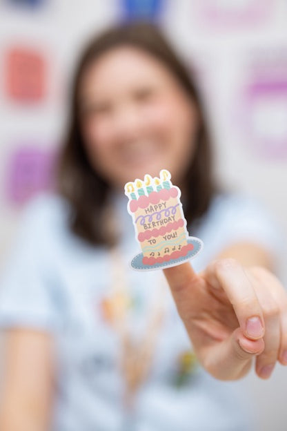 Person holding a small birthday cake sticker with colorful candles against a blurred background