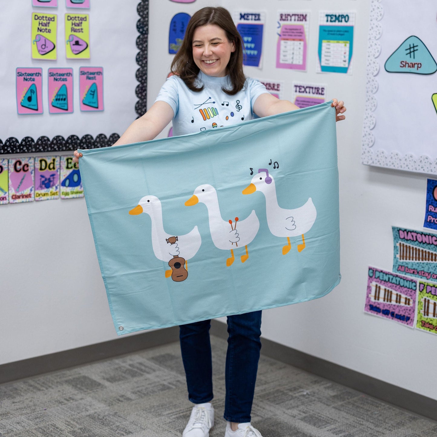 A woman in a classroom setting holds up a light blue fabric banner featuring three cartoon ducks playing musical instruments.