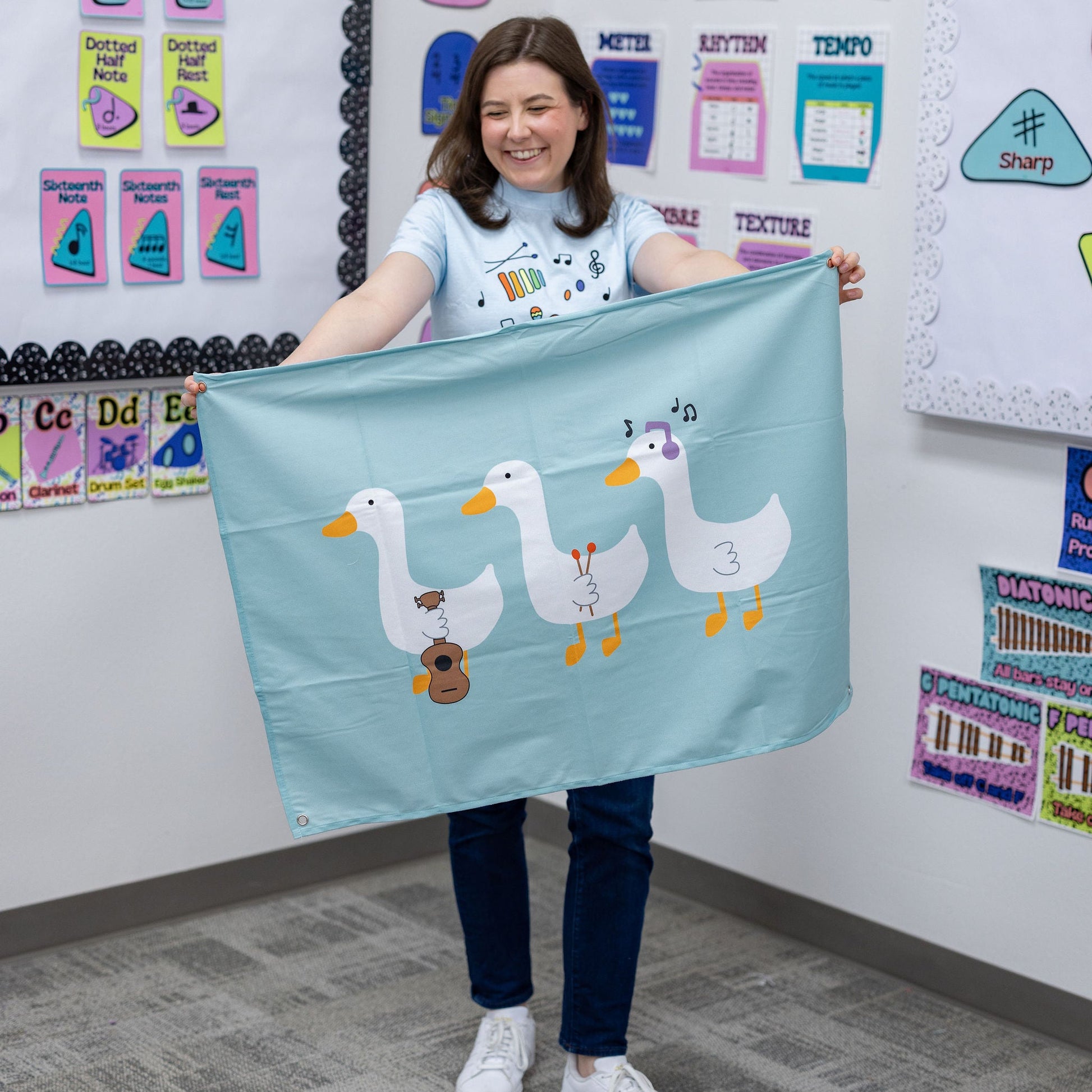 A woman in a classroom setting holds up a light blue fabric banner featuring three cartoon ducks playing musical instruments.